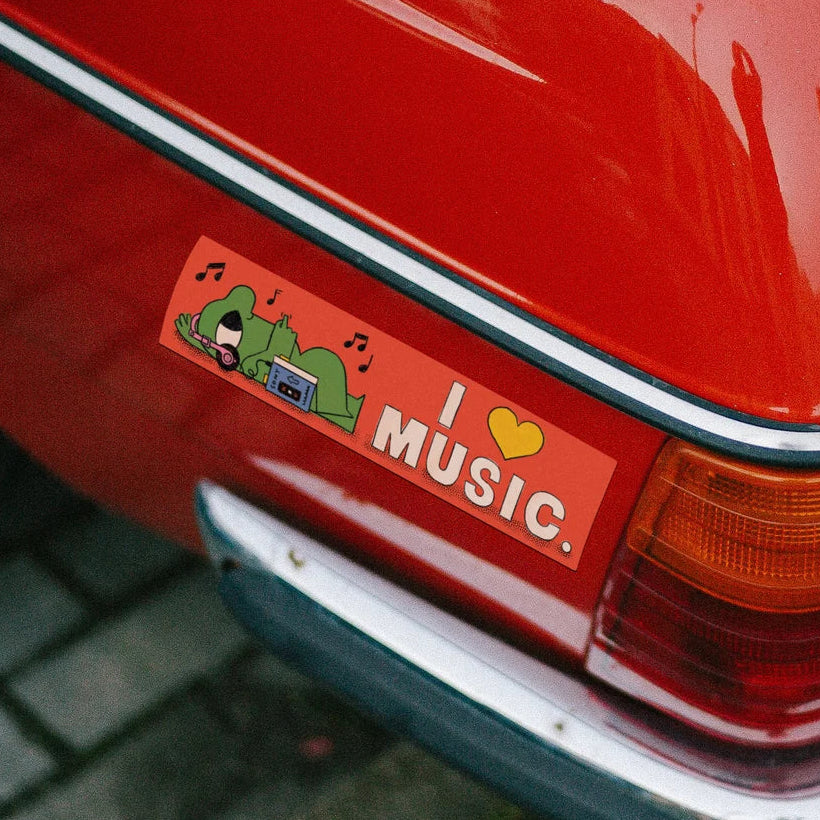 A close-up of a red car rear quarter panel showing a rectangular bumper sticker that reads 'I [heart] MUSIC.' with a small green character and cassette graphic, parked outdoors near pavement.