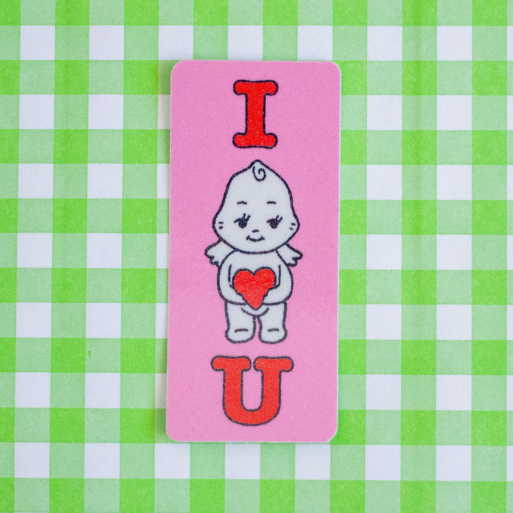 A rectangular pink sticker featuring a cute cartoon baby character holding an orange heart, with the letters 'I' and 'U' in orange above and below, placed on a green and white checkered background.