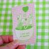 A hand holding a small rectangular sticker with a pink and white gingham background and green line art of a bunny, bear, hearts, and a matcha bowl. The card reads 'i love you so matcha' and is photographed against a green-and-white checkered surface.