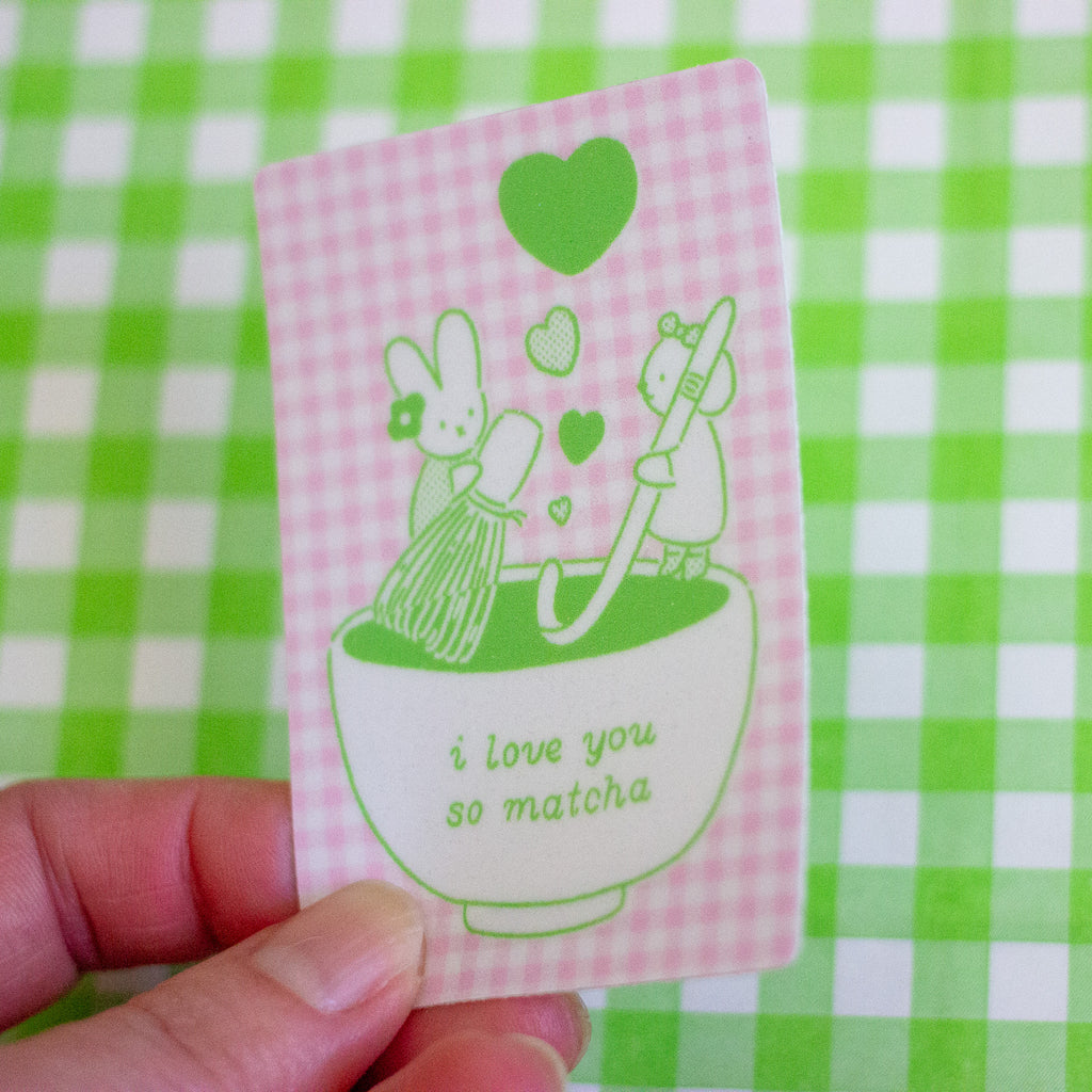 A hand holding a small rectangular sticker with a pink and white gingham background and green line art of a bunny, bear, hearts, and a matcha bowl. The card reads 'i love you so matcha' and is photographed against a green-and-white checkered surface.