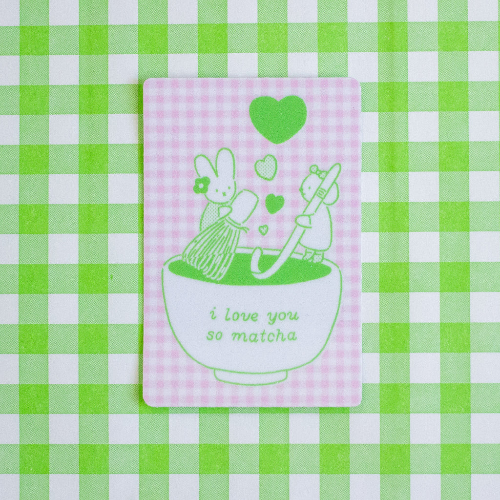 A rectangular pink and white gingham sticker featuring a green line-art illustration of two rabbits in a bowl with hearts and the text 'i love you so matcha', placed on a green and white checkered background.