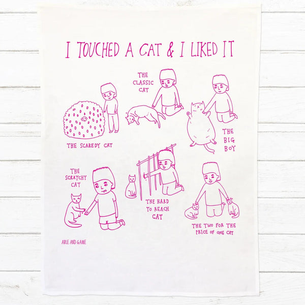 A white kitchen tea towel with pink line art illustrations and text, displayed flat on a white wood background.