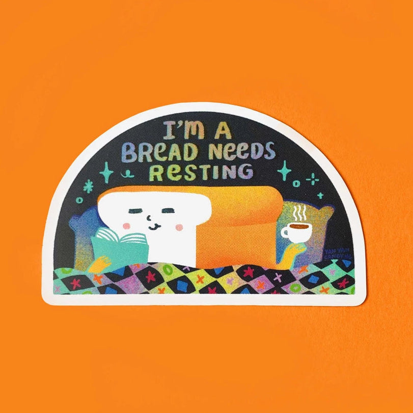 A die-cut sticker featuring a white slice of bread reading 'I'M A BREAD NEEDS RESTING' on a dark background, with a yellow-orange couch, a blue book, and a coffee cup on a colourful quilt, shown on a solid orange background.