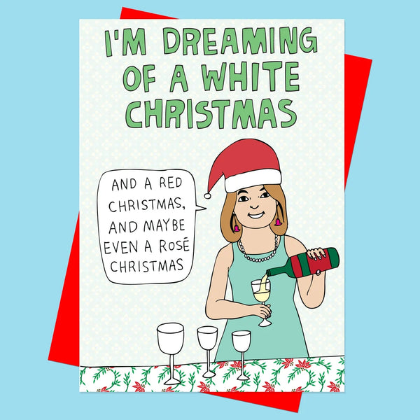 A Christmas greeting card shown against a light blue background, with a red envelope peeking out from behind it. The card features the text 'I'M DREAMING OF A WHITE CHRISTMAS' and a cartoon woman in a Santa hat pouring rosé wine, with a speech bubble reading 'AND A RED CHRISTMAS, AND MAYBE EVEN A ROSÉ CHRISTMAS'.