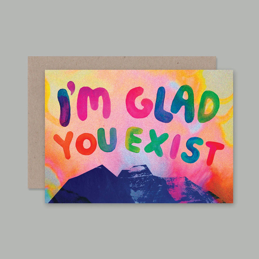 A greeting card with a kraft envelope, featuring the text 'I'M GLAD YOU EXIST' in colourful bubble letters over a watercolour-style background with yellow, pink, orange, blue, and purple tones, plus a dark blue mountain silhouette at the bottom.