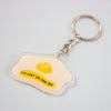 An acrylic keychain shaped like a fried egg, featuring a yellow yolk and the text 'I'M JUST AN EGG BOI' in black on a pale yellow background, attached to a silver split ring and chain.