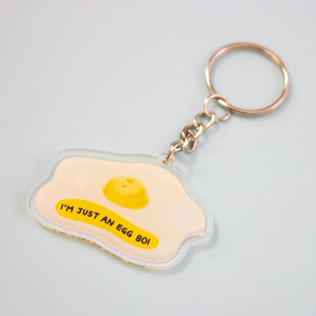 An acrylic keychain shaped like a fried egg, featuring a yellow yolk and the text 'I'M JUST AN EGG BOI' in black on a pale yellow background, attached to a silver split ring and chain.