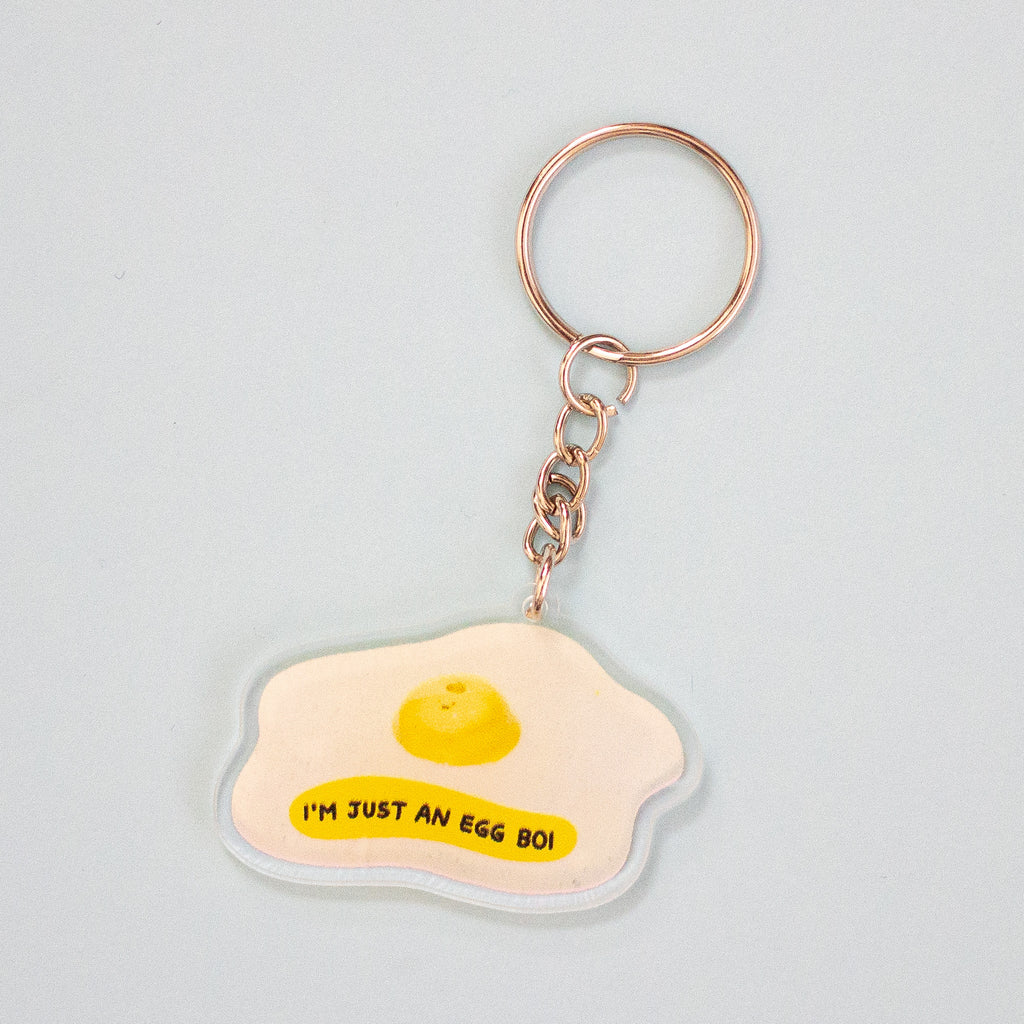 A keychain featuring a fried egg illustration with the text 'I'M JUST AN EGG BOI' on a light blue background, attached to a silver keyring with a key.