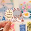 A hand holds a cute die-cut sticker featuring a grey cat writing in a notebook with the text 'it doesn't have to be good at all!' in a cream-coloured oval. The background shows a softly blurred, colourful classroom or desk scene with posters and notes pinned on a white board.