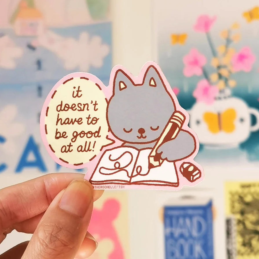 A hand holds a cute die-cut sticker featuring a grey cat writing in a notebook with the text 'it doesn't have to be good at all!' in a cream-coloured oval. The background shows a softly blurred, colourful classroom or desk scene with posters and notes pinned on a white board.