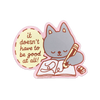 A die-cut sticker featuring a cute grey cartoon cat reading a book and writing in a pencil, with a pink speech bubble that says, 'it doesn't have to be good at all!'.