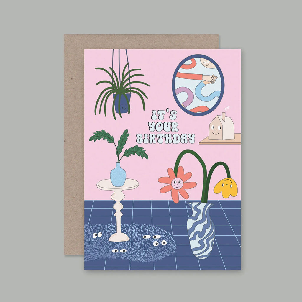 A birthday greeting card with a kraft envelope behind it, featuring a pastel illustration of a room with plants, flowers, a mirror, and the text 'IT'S YOUR BIRTHDAY' on a pink background.
