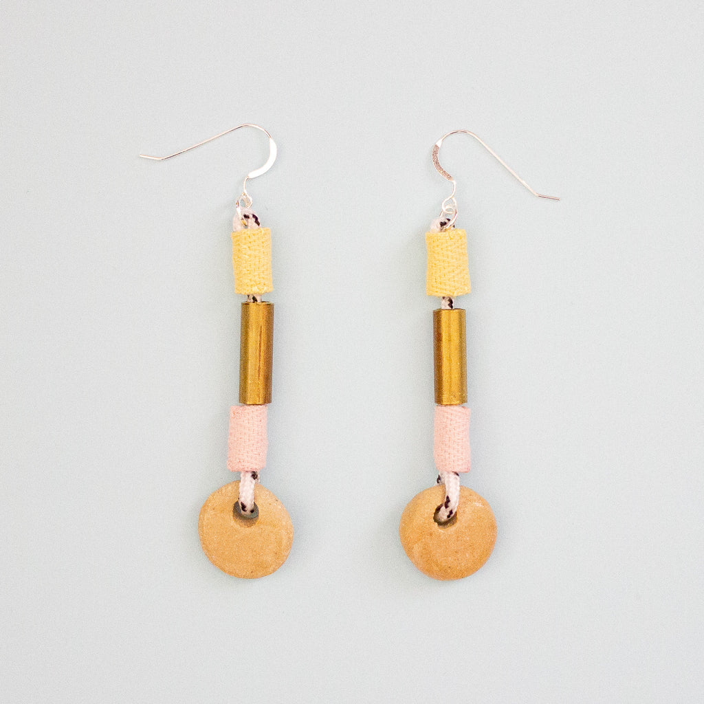 Pair of earrings with brass and pink and yellow fabric highlights, and ceramic beads on a light gray background