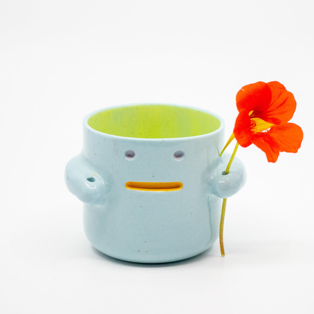 Ceramic monster-shaped tumbler with a yellow interior holding an orange flower on a white background