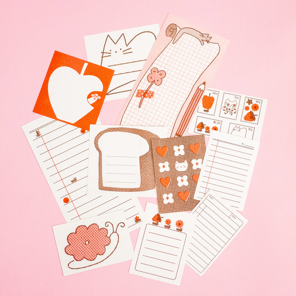 A flat-lay of cute journaling stationery in red, pink and brown tones, featuring designs of notepad pages, a pencil, a flower and a bread slice on a pink background.