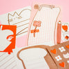 A styled flat-lay of cute stationery items on a pink background, including a pink grid notepad with a flower illustration, a white notepad with a cat outline, an orange card, a pencil, stamps, and patterned paper sheets.