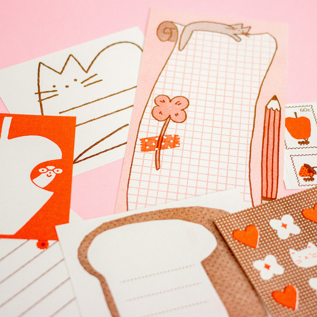 A styled flat-lay of cute stationery items on a pink background, including a pink grid notepad with a flower illustration, a white notepad with a cat outline, an orange card, a pencil, stamps, and patterned paper sheets.