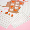 A flat-lay of cute journaling stationery in red, pink and brown tones, featuring designs of notepad pages, a pencil, a flower and a bread slice on a pink background.
