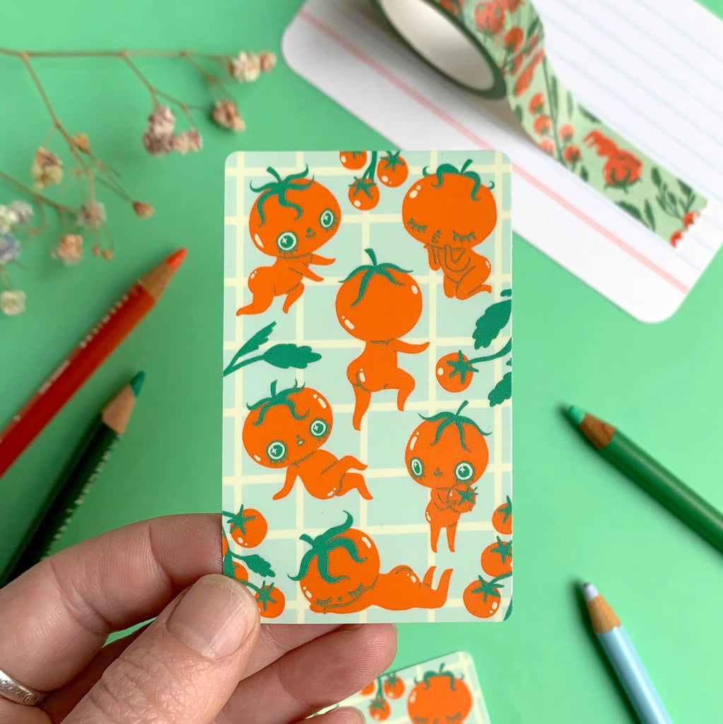 A hand holds a rectangular sticker featuring a playful pattern of cartoon tomatoes on a pale green grid background, with a matching roll of decorative tape visible in the upper right on a bright green surface surrounded by coloured pencils.