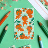 A flat-lay of patterned sticker featuring cute cartoon tomato characters on a mint green grid background, surrounded by coloured pencils and dried flowers on a mint green surface.