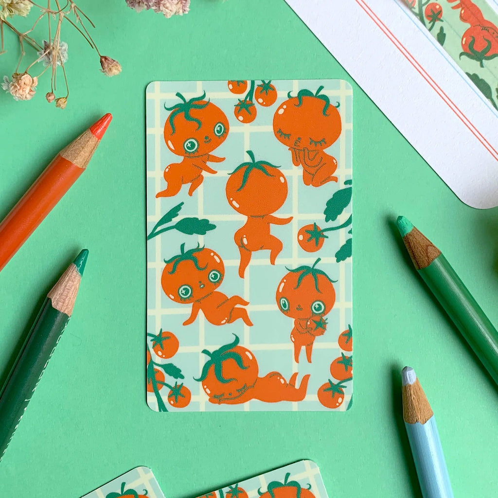A flat-lay of patterned sticker featuring cute cartoon tomato characters on a mint green grid background, surrounded by coloured pencils and dried flowers on a mint green surface.