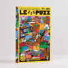 A boxed puzzle titled 'LE PUZZ' with a bright yellow border and a highly detailed, colourful cartoon illustration on the front showing many characters and objects. The box is photographed upright on a clean light grey studio background.