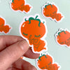 Hand holding a cute orange character sticker with more stickers in the background on a light mint surface.