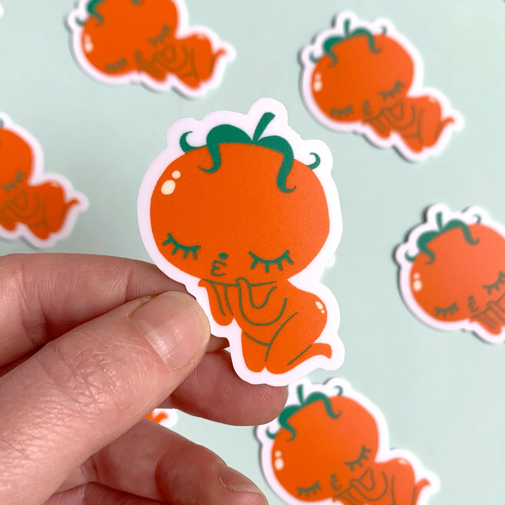 Hand holding a cute orange character sticker with more stickers in the background on a light mint surface.