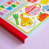 Side profile of a cute and colourful cover of a colouring postcard book against a lilac background.
