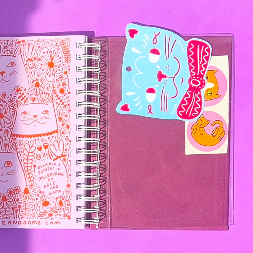 An open spiral notebook on a purple background, showing a printed page with orange cat illustrations and a pocket holding colorful cat-themed stickers.