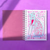 A spiral-bound notebook or art journal lies open on a purple surface. The left page is a solid dusty rose/magenta, and the right page features a white background with a colourful illustration of a cat in pink, light blue, and purple patterns.