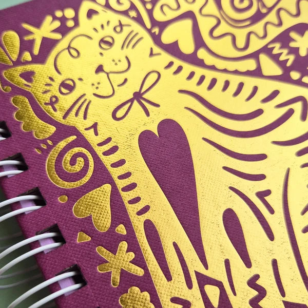 A close-up of a spiral-bound notebook with a textured purple cover decorated with shiny gold foil cat and heart illustrations.