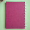 A magenta pink textured spiral notebook with a silver wire binding, shown flat on a light green background.