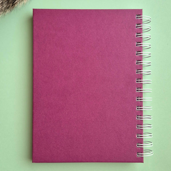 A magenta pink textured spiral notebook with a silver wire binding, shown flat on a light green background.