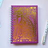 A purple spiral-bound notebook with a metallic gold foil cat design on the cover, styled on a light pink background with a pen, binder clip, and patterned paper.