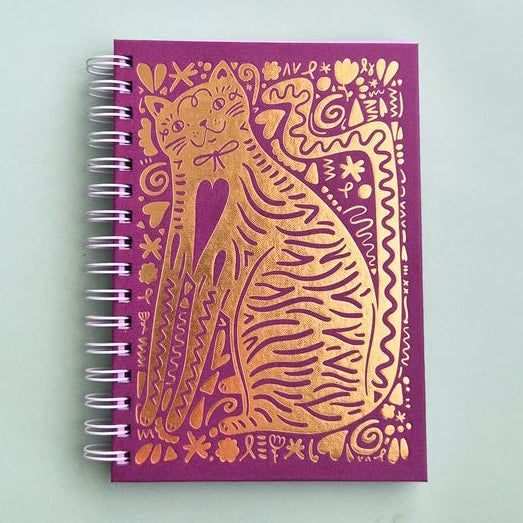 A purple spiral-bound notebook featuring a large gold foil illustration of a cat on the cover, photographed on a light mint green background.