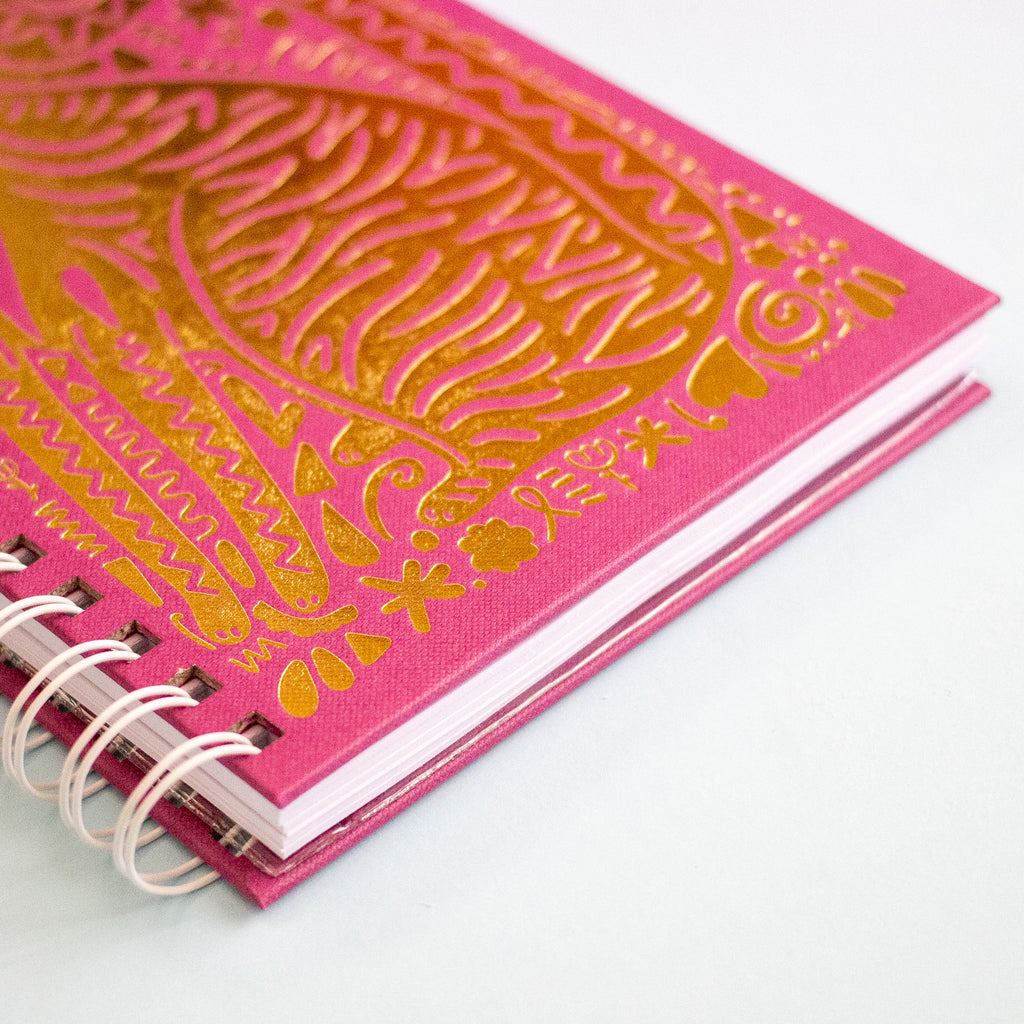 A close-up of a purple spiral-bound notebook with metallic gold foil embossed patterns on the cover, photographed on a clean white background.