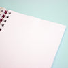A close-up of an open spiral-bound notebook with a blank, lightly dotted page on a pastel aqua background.