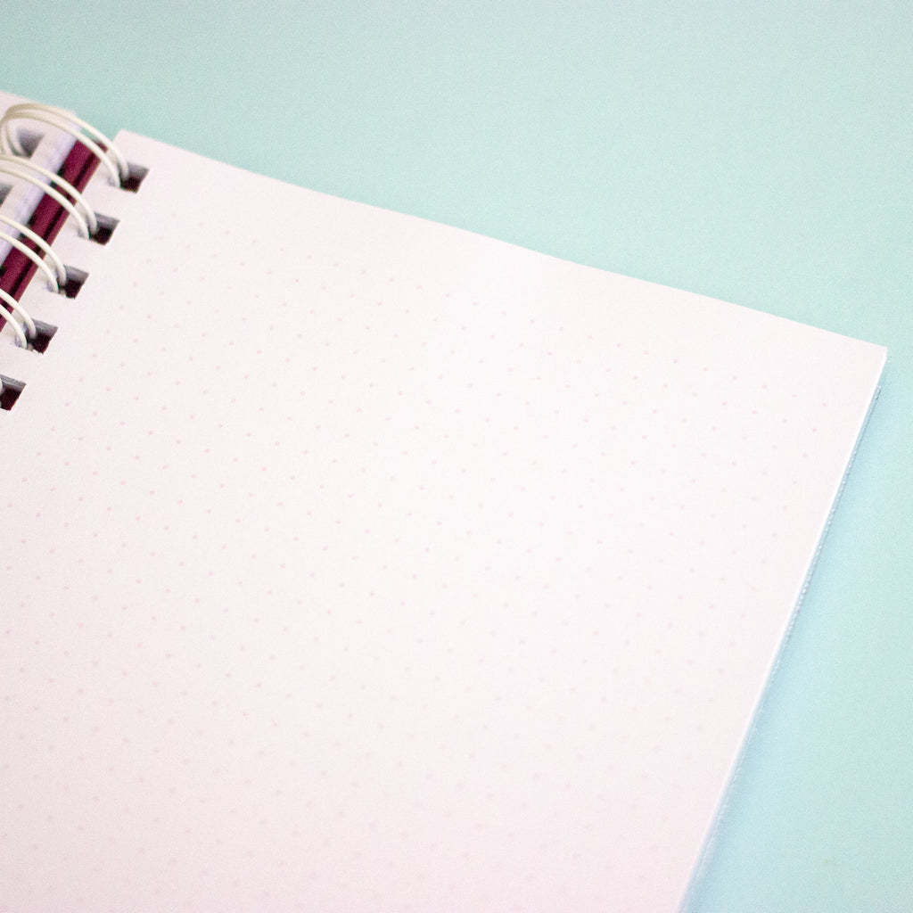 A close-up of an open spiral-bound notebook with a blank, lightly dotted page on a pastel aqua background.