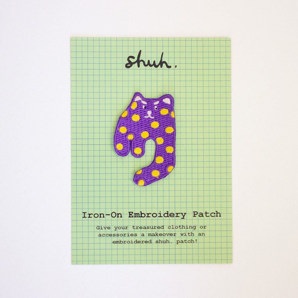 Purple cat-shaped iron-on embroidery patch with yellow dots on a green grid card.