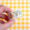 A hand holding a roll of washi tape with a floral and bunny pattern, set against a yellow and white checkered background.