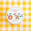 A roll of Kusamura tape by Aiko Fukawa, featuring a white label with a watercolour rabbit and floral illustrations, placed on a yellow and white checkered background.