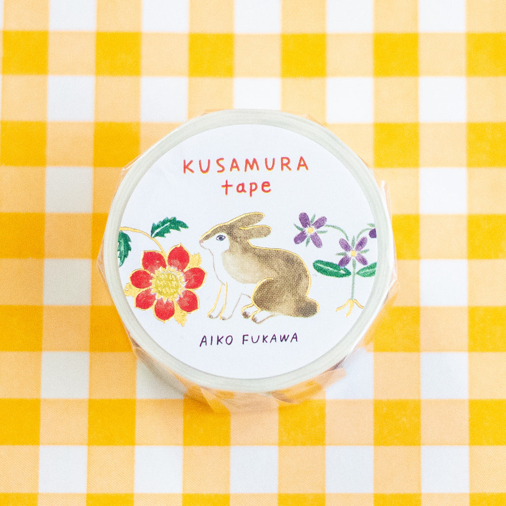 A roll of Kusamura tape by Aiko Fukawa, featuring a white label with a watercolour rabbit and floral illustrations, placed on a yellow and white checkered background.