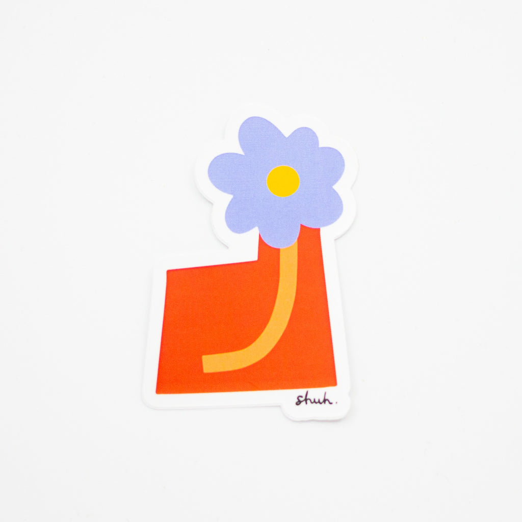 A purple flower, orange stem, in a red vase. Vase is square shaped, offset like the letter L. 