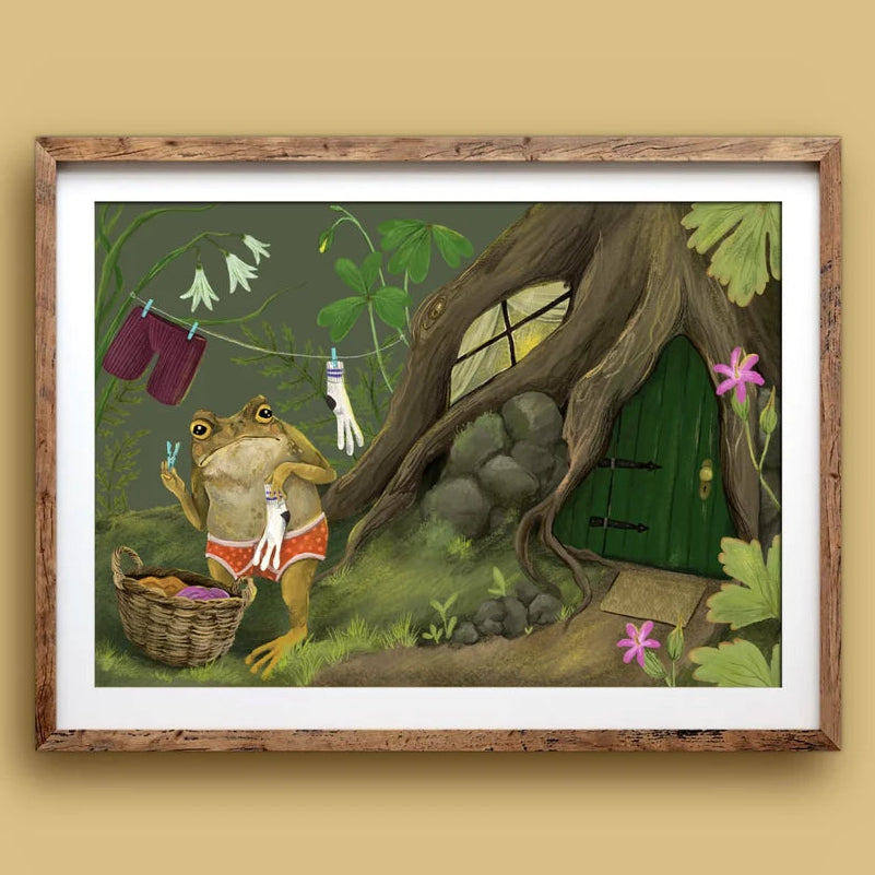A framed art print featuring a whimsical illustration of a frog washing its clothes in a forest setting with a treehouse door, mounted on a beige wall.