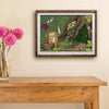 A framed art print depicting a whimsical frog character doing laundry, styled on a beige wall above a wooden console table with a vase of pink flowers.