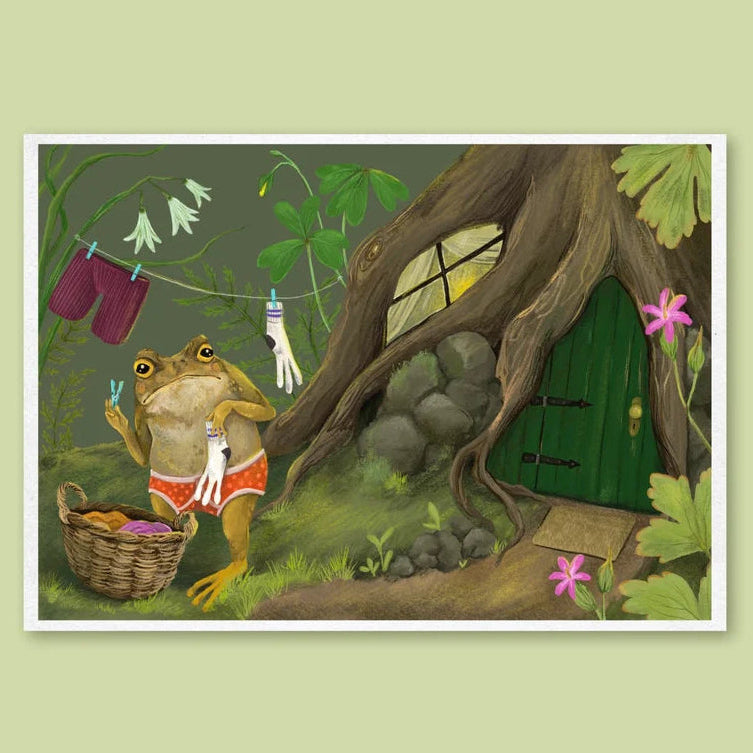 A framed art print featuring a whimsical illustration of a frog washing clothes outside a treehouse, set against a solid pale green background.