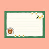 A square lined writing card with a green border, cream center, small white flowers, and a teddy bear illustration in a teacup at the bottom left, shown on a peach background.
