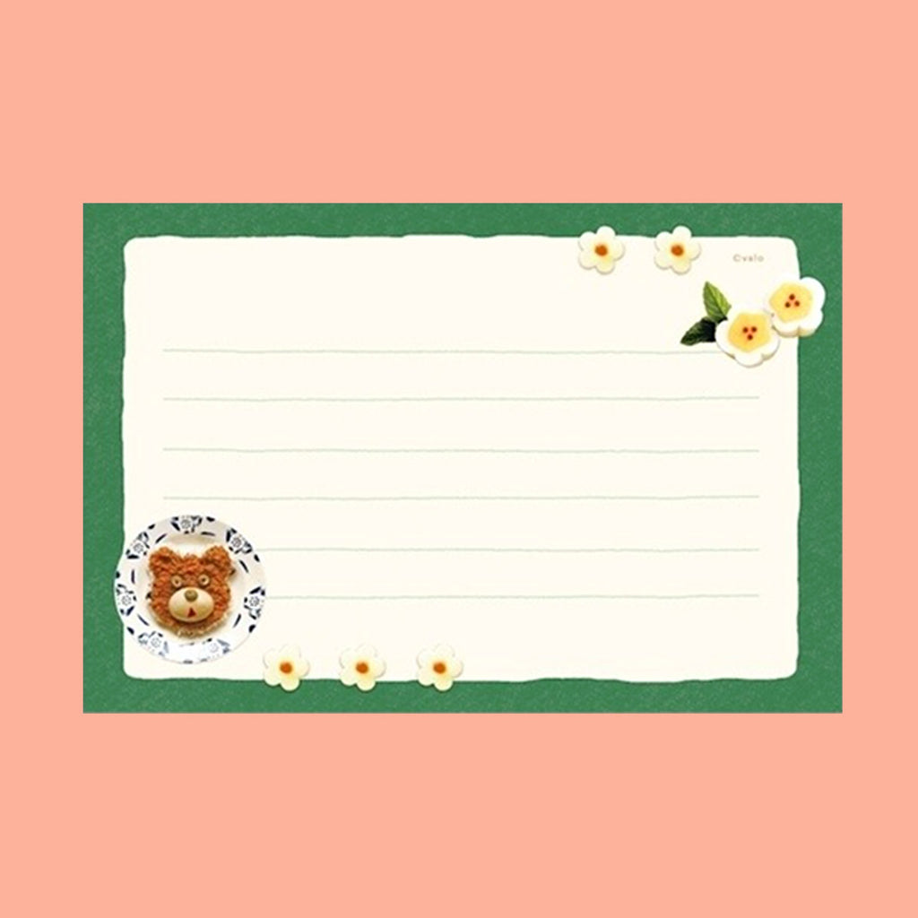 A square lined writing card with a green border, cream center, small white flowers, and a teddy bear illustration in a teacup at the bottom left, shown on a peach background.