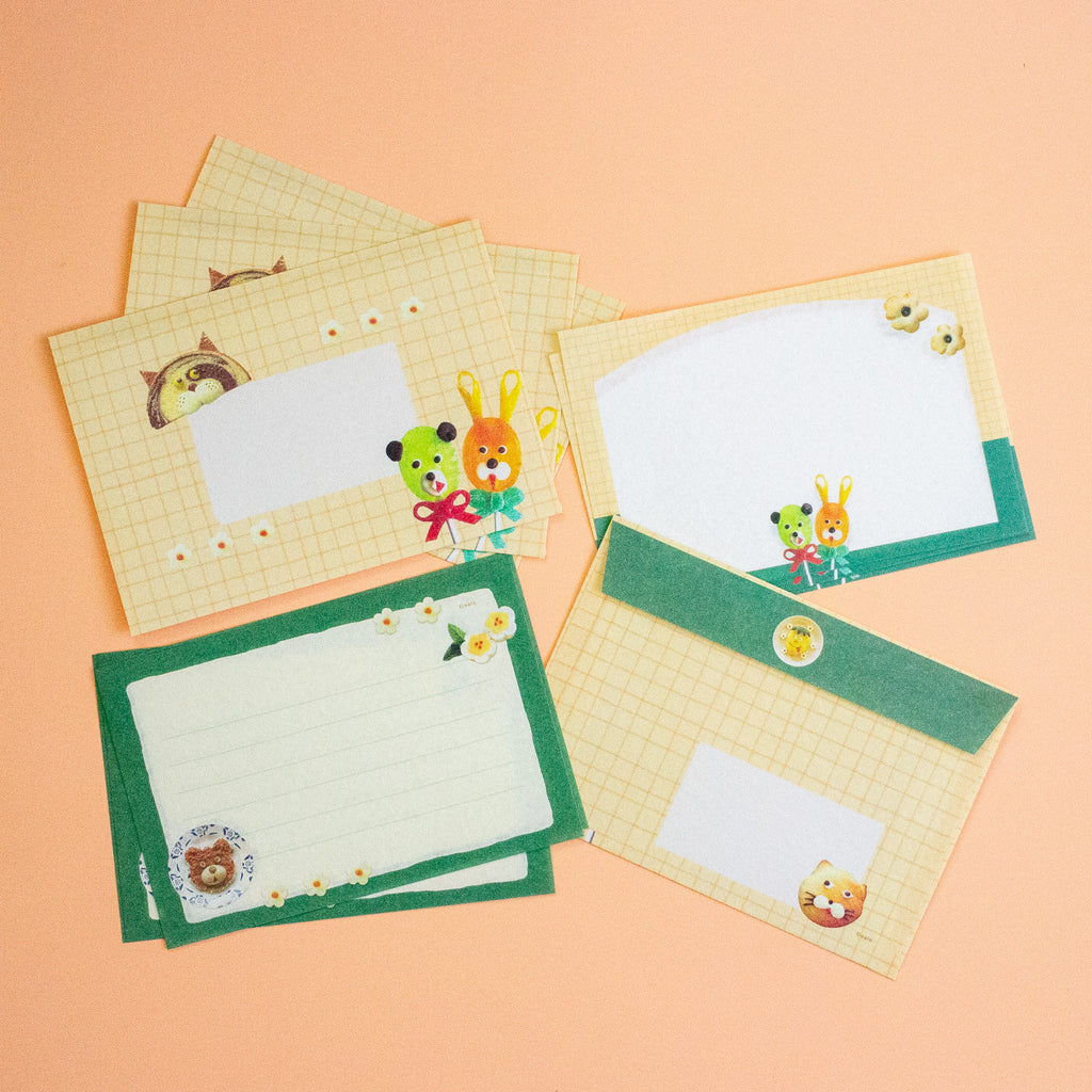 A set of cute stationery cards and envelopes with green borders, a yellow grid pattern, and cartoon animal illustrations, arranged on a peach-coloured background.
